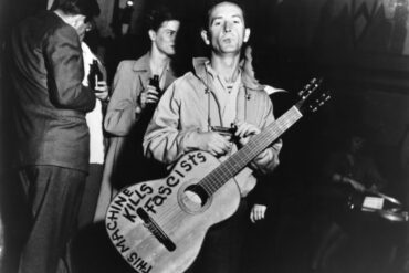 Woody Guthrie, standing, with a guitar around his neck and the written message, "This Machine Kills Fascists" © Lester Balog
