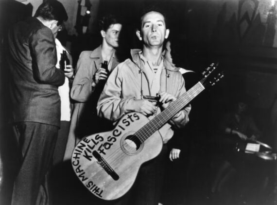 Woody Guthrie, standing, with a guitar around his neck and the written message, "This Machine Kills Fascists" © Lester Balog